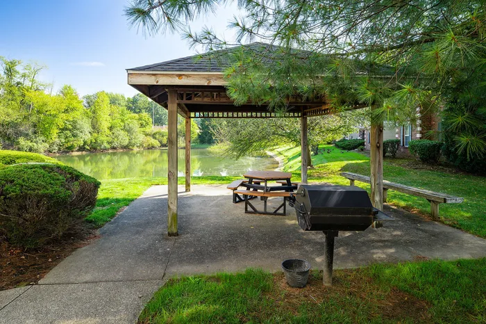 Charming outdoor gazebo with picnic table overlooking a tranquil pond, surrounded by lush greenery and featuring a convenient barbecue grill.