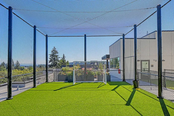 Inviting rooftop mini soccer field with netted enclosure, lush green turf, and a panoramic city view, perfect for active living.