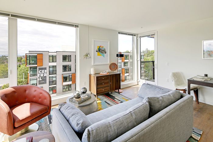 Bright and inviting living room with modern decor, featuring a cozy armchair, plush gray sofa, and large windows with a view of a stylish apartment complex.