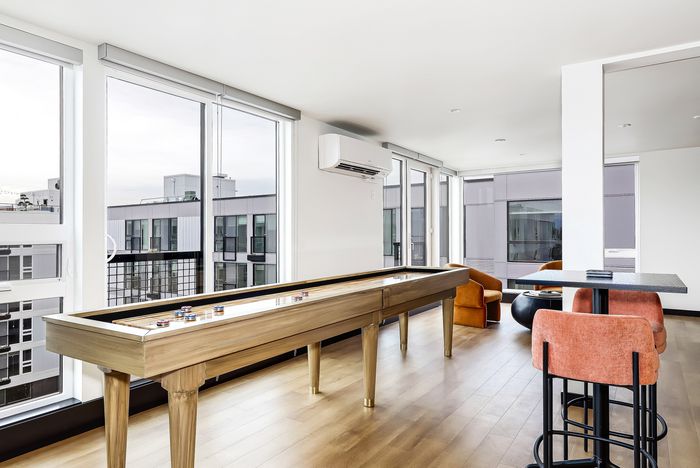 Bright modern apartment with expansive windows, chic orange seating, shuffleboard table, and hardwood floors, offering stylish urban views.