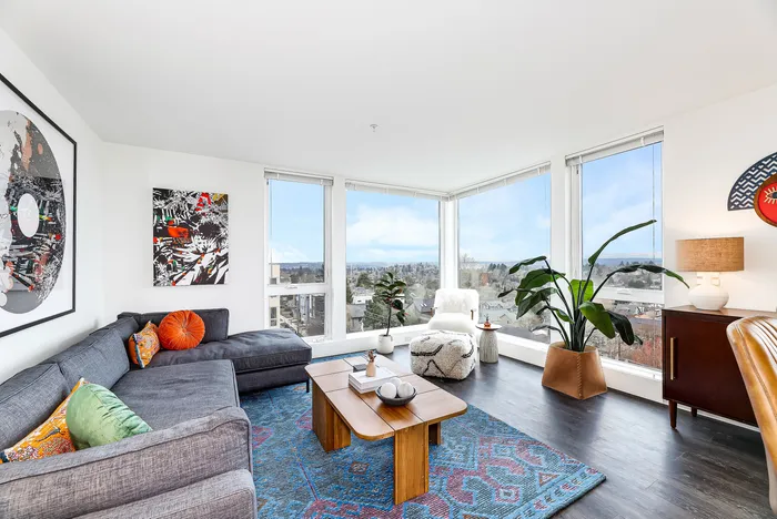 Bright and modern living room with large windows offering scenic city views, stylish gray sectional sofa, vibrant artwork, and lush indoor plants.
