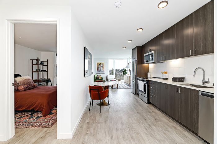 Modern open-plan apartment kitchen with sleek dark wood cabinetry, adjacent cozy living area with vibrant seating, and a glimpse into a stylishly furnished bedroom.