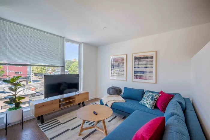 Bright and modern living room featuring a stylish blue sectional sofa, contemporary art, and a sleek TV setup with large windows showcasing natural light.