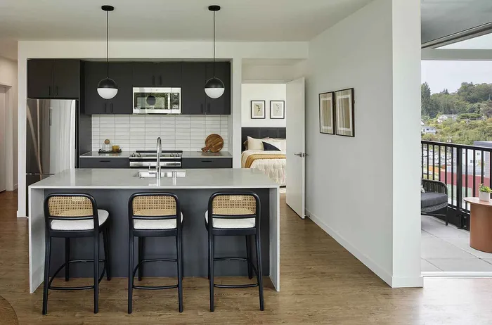 Modern apartment kitchen featuring sleek cabinetry, a stylish island with seating, and a view of a cozy bedroom and balcony.