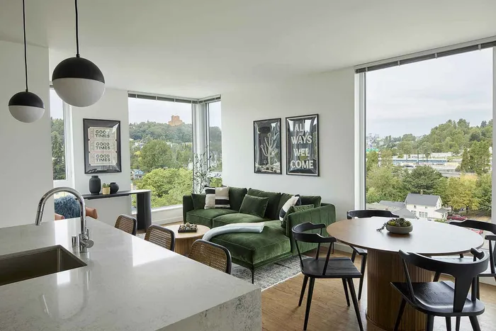 Modern living area with large windows offering stunning views, a plush green sofa, stylish artwork, and a cozy dining space in an open-concept apartment.