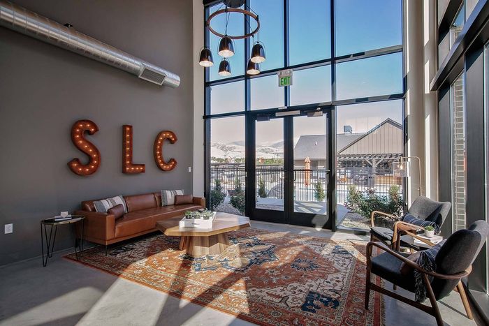 Modern lounge area featuring a cozy leather sofa, stylish chairs, and a chic coffee table with a scenic view through large glass doors. The room is elegantly accented by an artistic 'SLC' wall decoration and an intricate red and blue patterned rug.