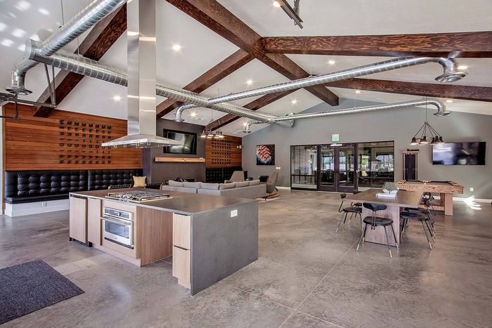 Modern open-concept community space with industrial-style kitchen, cozy lounge area, and stylish communal table, featuring exposed beams and ductwork.