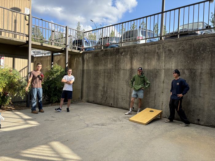 Sunny outdoor area with people enjoying a game of cornhole, surrounded by greenery and urban architecture.