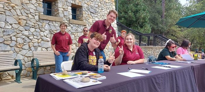 Enthusiastic group of people at an outdoor information table, with volunteers in maroon shirts ready to assist in a welcoming setting.