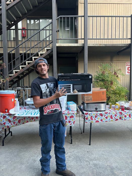 Person standing proudly in front of an apartment complex, holding a new countertop microwave oven. A festive table set with food and drinks adds to a welcoming community vibe.