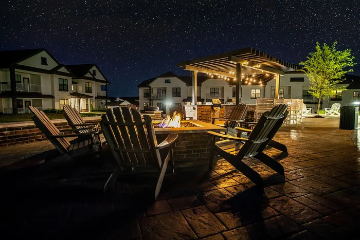 Enchanting nighttime view of a cozy outdoor fire pit area with Adirondack chairs, pergola, and twinkling string lights under a starry sky, surrounded by modern apartment buildings.
