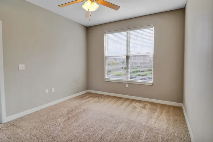 Bright apartment bedroom featuring plush carpeting, large window with city views, and elegant ceiling fan.