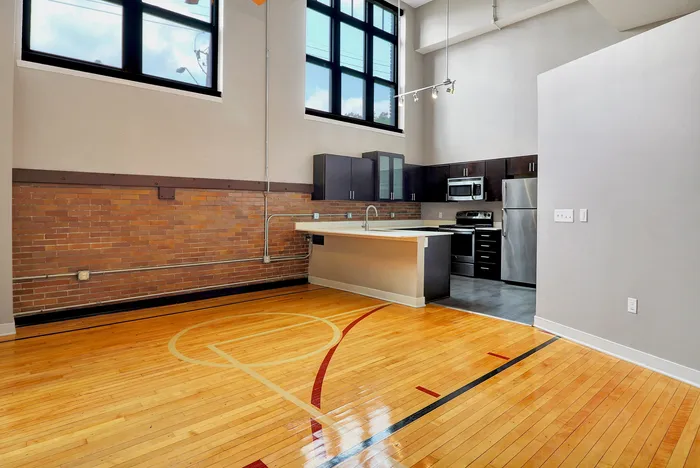 Loft-style kitchen with modern appliances and high ceilings, adjoining a unique hardwood basketball floor.