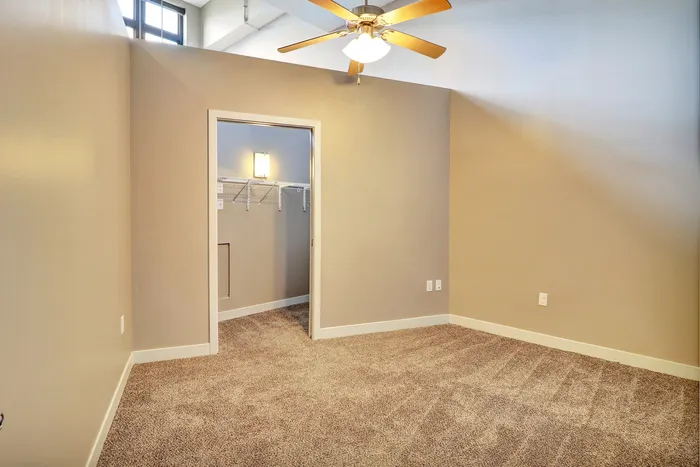 Inviting bedroom with plush carpeting and walk-in closet, illuminated by a ceiling fan and natural light from upper windows.