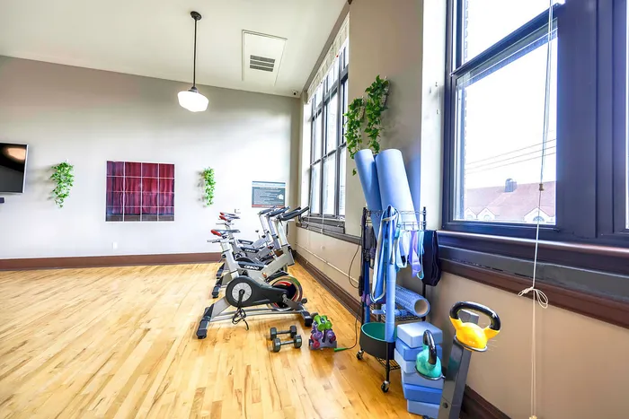 Bright and inviting gym space featuring stationary bikes, yoga mats, and modern fitness equipment, enhanced by large windows and elegant wood flooring.