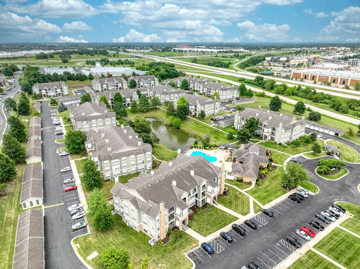 Aerial view of a modern apartment complex with abundant green space, a central pond with fountain, and a sparkling community pool, offering serene suburban living.