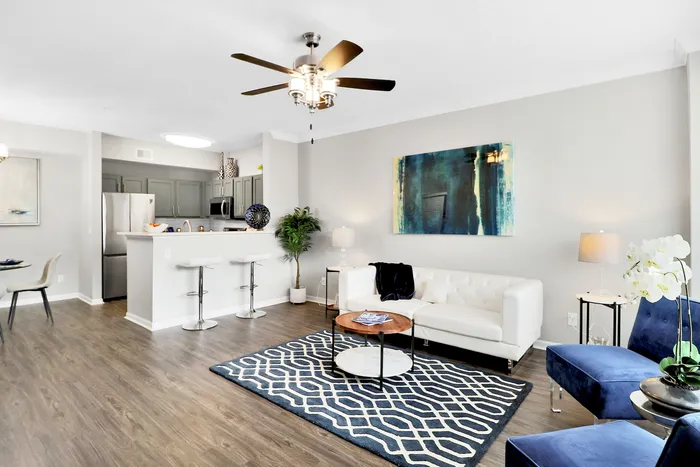 Modern living room with elegant wood flooring, featuring a white sofa, vibrant blue chairs, and a contemporary kitchen with stainless steel appliances.