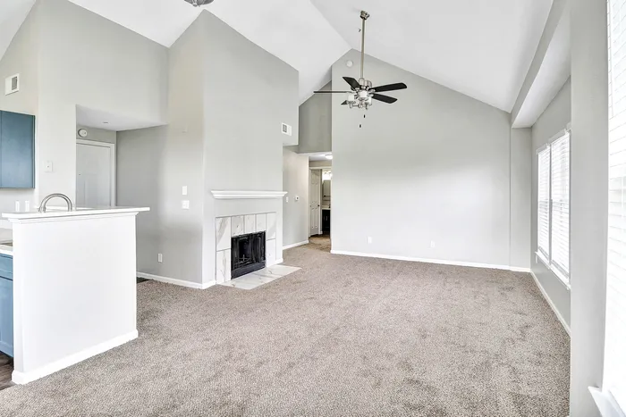 Bright and airy living room with vaulted ceilings, modern ceiling fan, and a cozy fireplace, featuring a plush carpet and ample natural light from large windows.