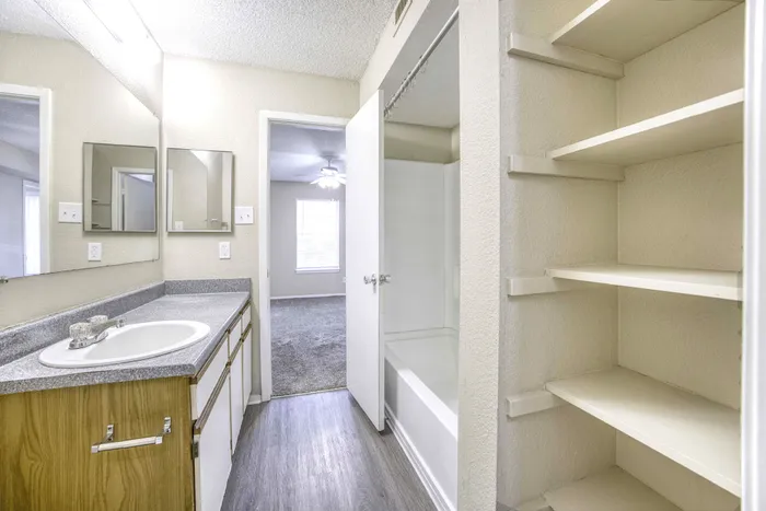 Bright, modern bathroom with ample shelving, sleek countertop, and an inviting tub adjoining a cozy carpeted room with natural light.
