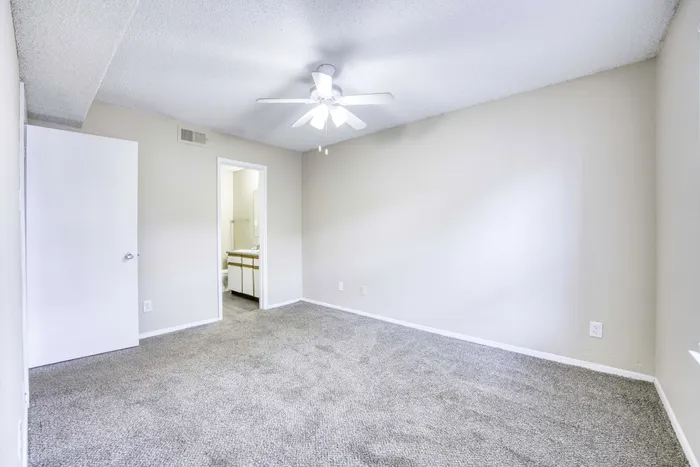 Bright and inviting bedroom with soft carpeting, neutral walls, and a modern ceiling fan, leading into a well-lit bathroom.