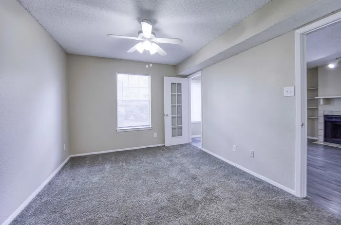 Bright and spacious room with fresh carpeting, modern ceiling fan, and a door leading to a cozy living space with a fireplace.
