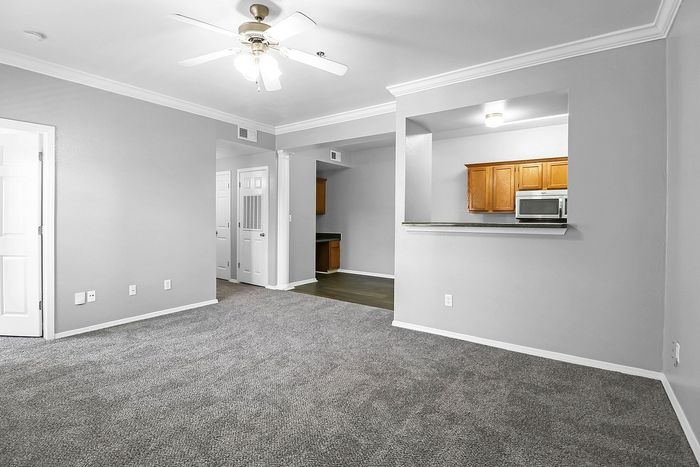 Modern apartment living area with plush carpeting, ceiling fan, and a sleek pass-through kitchen.