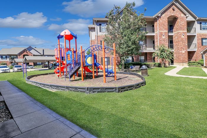 Charming apartment complex with vibrant playground, lush green lawn, and stylish brick buildings under a clear blue sky.