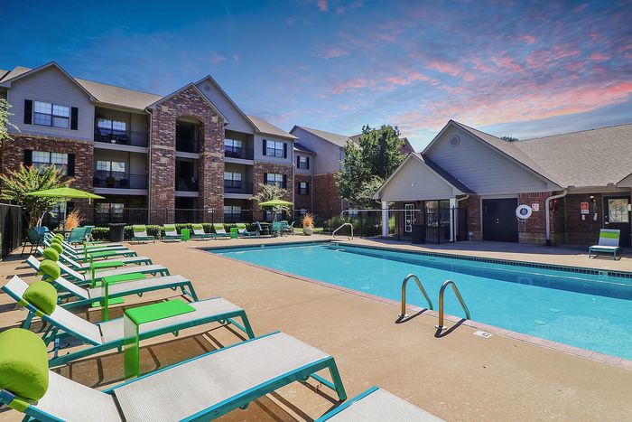 Inviting apartment complex with a sparkling swimming pool, surrounded by modern lounge chairs and lush greenery under a picturesque sky.