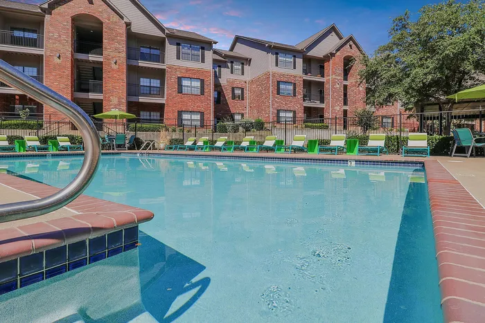 Luxurious apartment complex with a sparkling swimming pool, surrounded by vibrant green lounge chairs and beautiful brick architecture.