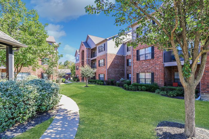 Charming brick apartment complex surrounded by lush greenery and a well-maintained garden path under a clear blue sky.
