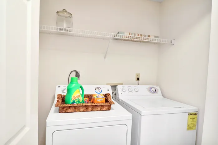 Modern laundry room with a washer and dryer, featuring a convenient shelf for storage and a tidy basket with cleaning supplies.