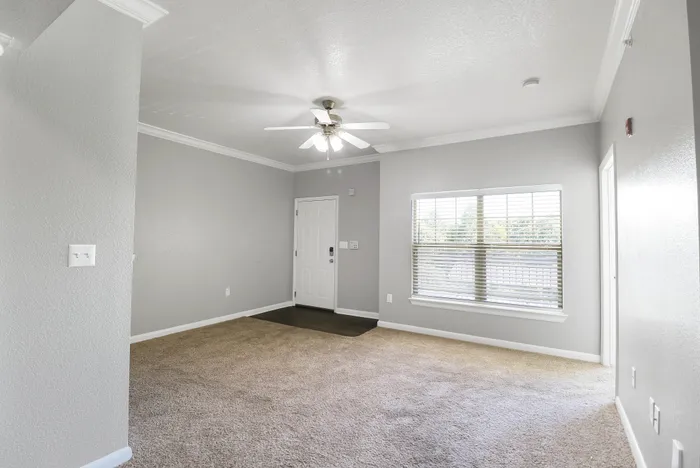 Bright, spacious living room with neutral carpet, ceiling fan, and large window for natural light.