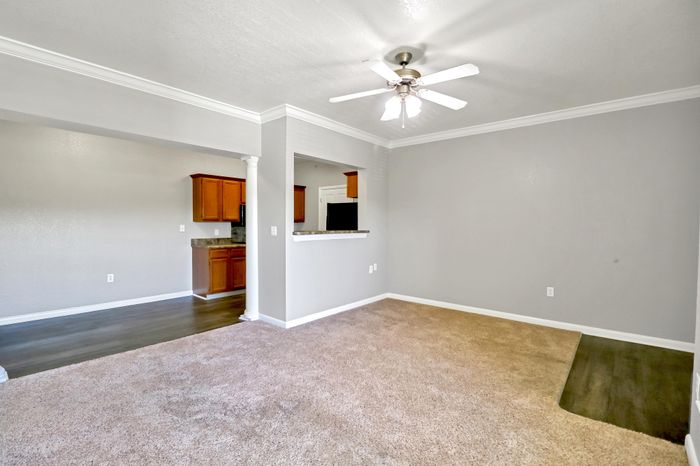 Inviting living area with plush carpeting, crown molding, and a ceiling fan, adjoining a modern kitchen with rich wood cabinetry.