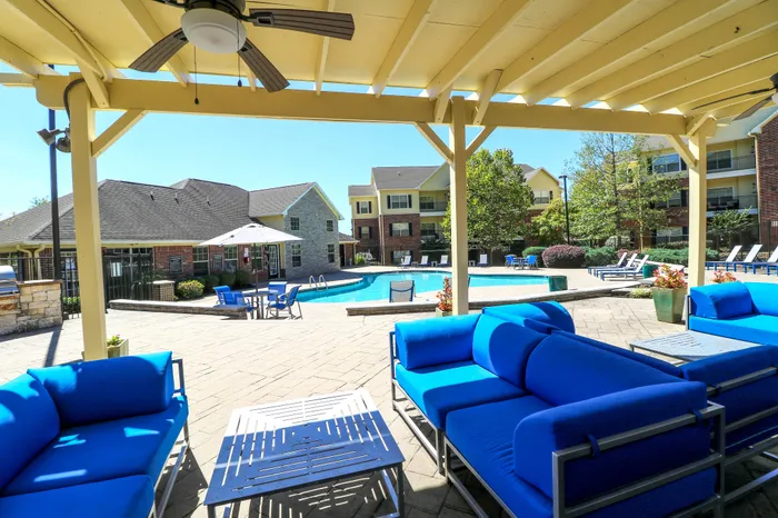 Inviting apartment complex with modern blue seating under a pergola, overlooking a sparkling pool surrounded by lush greenery and stylish buildings.