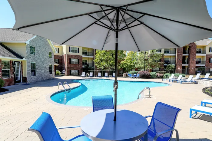 Inviting outdoor pool area with modern seating and umbrella, surrounded by landscaped gardens and attractive brick and stone apartment buildings.