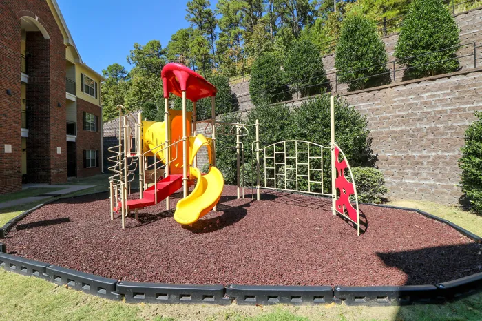 Charming apartment complex playground with vibrant yellow and red slides, surrounded by lush greenery and brick buildings.