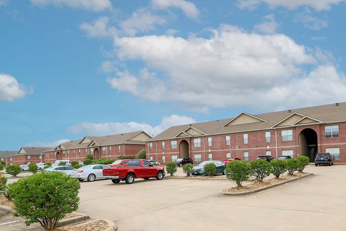 Charming brick apartment complex with ample parking and lush greenery under a bright blue sky.