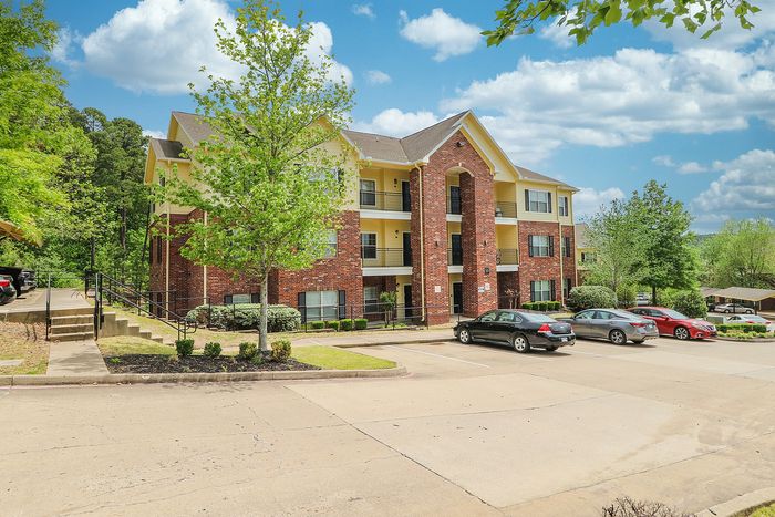 Inviting apartment complex with brick facade, surrounded by lush greenery and clear blue skies, offering spacious, modern living.