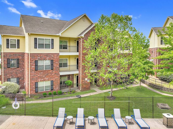 Charming multi-story apartment building with stylish brick facade and lush greenery, featuring a refreshing outdoor pool area with sun loungers for relaxation.