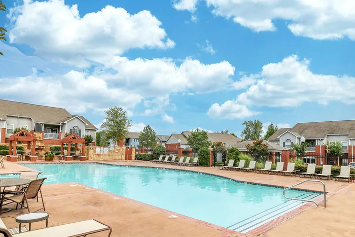 Inviting and sunny community pool area with comfortable lounge chairs, surrounded by charming residential buildings and lush greenery under a vibrant blue sky.