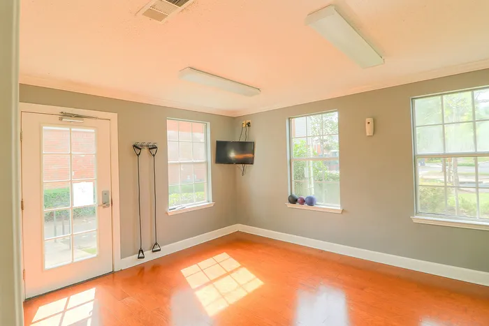 Bright and inviting exercise room with wooden floors, a wall-mounted TV, and fitness equipment by large windows.