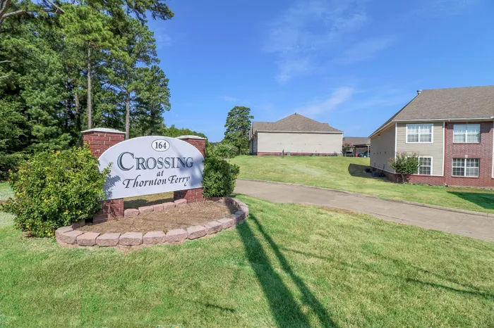 Inviting entrance to Crossing at Thornton Ferry, featuring lush green lawns and charming brick-accented buildings under a clear blue sky.