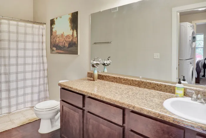 Inviting bathroom with elegant granite countertop, spacious mirror, and modern stacked washer and dryer in view, perfect for convenient living.