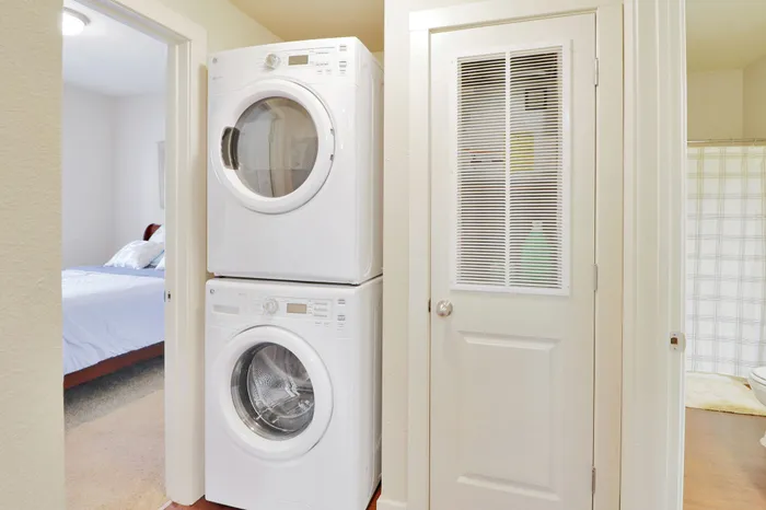 Bright apartment laundry area with modern stacked washer and dryer adjacent to cozy bedroom and bathroom.