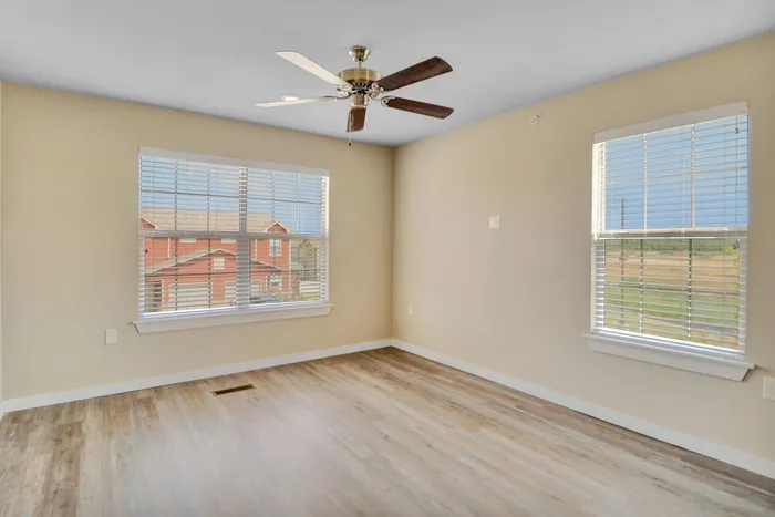 Bright and inviting room with stylish wood flooring, large windows with blinds, and a modern ceiling fan.