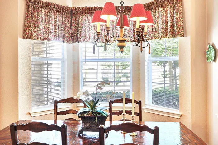 Elegant dining area with warm lighting from a charming chandelier, featuring a wooden table accented by a floral centerpiece and surrounded by large sunlit windows.