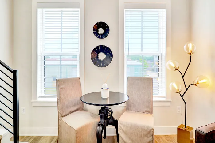 Chic and bright seating area with elegant round table, two stylish chairs, artistic wall decor, and modern floor lamp with globe bulbs near large windows.
