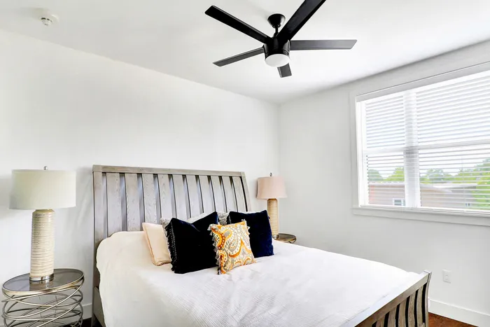 Inviting bedroom with modern ceiling fan, elegant wooden bed frame, and decorative pillows, bathed in natural light from a large window.