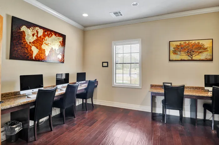 Elegant home office with four modern computers on a granite desk, complemented by artistic world map and tree paintings, featuring chic dark wood flooring and abundant natural light.