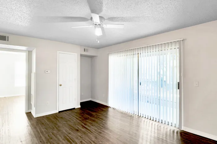 Bright living room featuring sleek wood flooring, ceiling fan, and large sliding glass doors with vertical blinds for ample natural light.